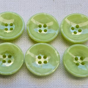 May include: Six light green, round buttons with four holes each. The buttons have a glossy finish and a slightly concave shape. The buttons are arranged in two rows of three.