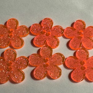 Peut inclure: Six boutons orange en forme de fleur avec des paillettes. Chaque bouton a cinq pétales et un petit trou au centre pour la couture. Les boutons sont en matériau translucide.