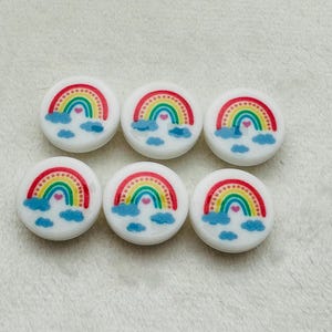 Puede incluir: Seis botones redondos blancos con un diseño de arcoíris y nubes. Cada botón tiene un arcoíris rojo, naranja, amarillo, verde, azul y morado con un pequeño corazón rosa en el centro. Las nubes son blancas con un contorno azul claro.