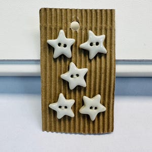 May include: Five off-white star-shaped buttons with two holes in the centre, arranged in a staggered pattern on a rectangular piece of brown corrugated cardboard with a circular hole at the top for hanging display purposes.