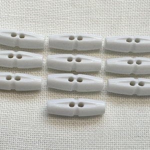 May include: A collection of ten white toggle buttons arranged in three rows. Each button is elongated with a smooth, glossy finish, featuring two centred holes for attachment and a slightly tapered shape at both ends for a classic fastener look.