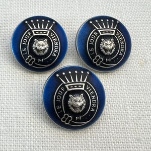 May include: Three circular buttons with a blue and black design. Each button features a silver lion's head, a crown, and the text "LE JOUR VIENDRA". These buttons are likely for clothing or craft projects.