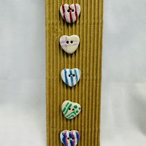 May include: Five heart-shaped ceramic buttons with colourful striped and dotted designs. The buttons are white with red, blue, green, and purple accents. They are attached to a corrugated cardboard backing.
