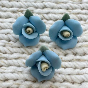 May include: Three small, sculpted flower ornaments in a light blue colour. Each flower features rounded, layered petals, a pale cream-coloured centre, and a single small, pointed green leaf at the top. The surface of the flowers appears smooth and matte.