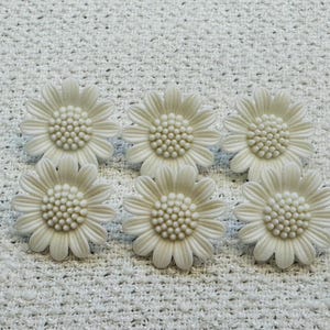 May include: Six off-white daisy-shaped decorative embellishments. Each flower has a detailed centre and petals, arranged on a textured white surface. The daisies are uniform in size and colour, creating a cohesive floral design.