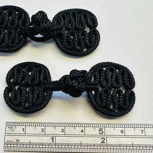 May include: Two black frog closures, a type of button, are displayed against a white background. Each closure is made of woven black cord and has a distinctive looped design. A ruler shows the closures are about 2 inches long.