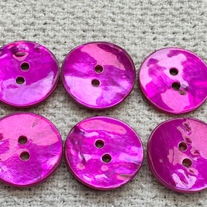 May include: Six round, iridescent magenta buttons with two or three holes each. The buttons have a shiny, reflective surface and are arranged in two rows of three on a textured, off-white background.