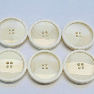 May include: Six large, round, off-white buttons with four holes each. The buttons have a slightly iridescent, pearlescent center and a smooth, white rim. They are arranged in two rows of three on a light background.