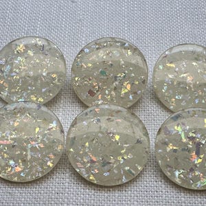 May include: Six round, iridescent buttons with a clear, glittery appearance. The buttons are a light, creamy colour and are scattered with colourful, reflective flakes. The buttons are arranged in two rows of three.