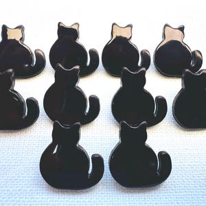 Puede incluir: Una colección de diez botones negros con forma de gato. Cada botón presenta un acabado brillante y un diseño sencillo de gato sentado. Los botones están dispuestos sobre una superficie azul claro.
