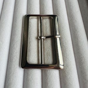 Puede incluir: Hebilla de cinturón rectangular plateada brillante con una sola barra y una espiga. La hebilla tiene un acabado pulido. El fondo es una superficie clara y texturizada.