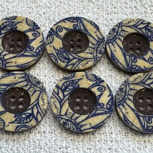 May include: Six round buttons with a cream background and a dark blue floral design. Each button has a dark brown centre with four holes for sewing. The buttons are arranged in two rows of three.