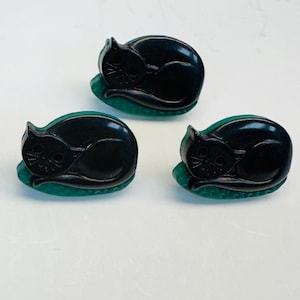 May include: Three black cat-shaped decorative objects, each resting on a small, green, oval base. The cats are in a curled-up sleeping position, with visible facial details. The objects are displayed against a white background.