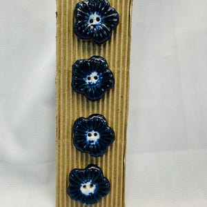 May include: Four dark blue ceramic flower-shaped buttons with white centres are displayed on a corrugated cardboard strip. The buttons have a glossy finish and are arranged vertically. The background is a plain white surface.