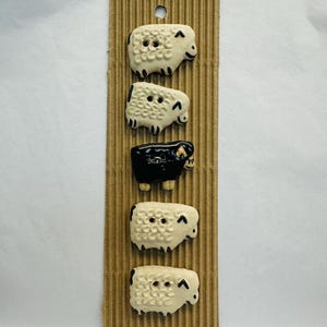 Puede incluir: Un conjunto de cinco botones con forma de oveja en crema y negro, sujetos a un respaldo de cartón corrugado. Los botones tienen dos o tres agujeros para coser. Las ovejas tienen un diseño de dibujos animados con pequeños ojos negros y una boca sonriente.