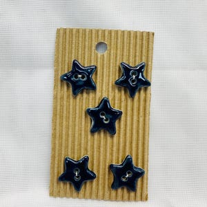 May include: Five navy blue star-shaped buttons with white detailing are presented on a corrugated cardboard backing. Each button features two holes for sewing. The buttons are arranged vertically.