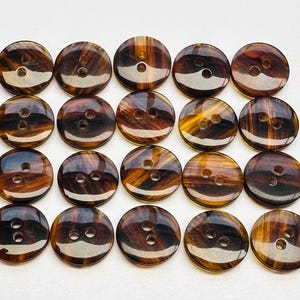 Puede incluir: Una colección de veintiún botones redondos con apariencia veteada. Los botones son principalmente marrones con vetas ámbar y marrón oscuro. Cada botón tiene dos agujeros para coser y un acabado brillante.