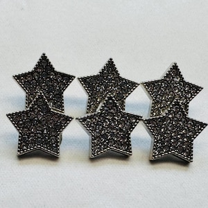 Puede incluir: Seis pendientes de botón con forma de estrella. Cada pendiente es plateado e incrustado con pequeños cristales de color gris oscuro. Los pendientes están dispuestos en un grupo sobre una superficie blanca.