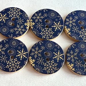 May include: Six round wooden buttons with a navy blue background and a gold snowflake pattern. Each button has two holes for sewing and a light wood edge. These buttons are ideal for winter or holiday-themed crafts.