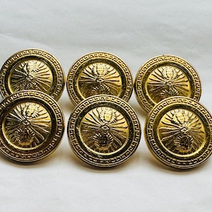 May include: Six gold-toned buttons with a raised design featuring a stylised sunburst and a face in the centre. The buttons have a decorative border.