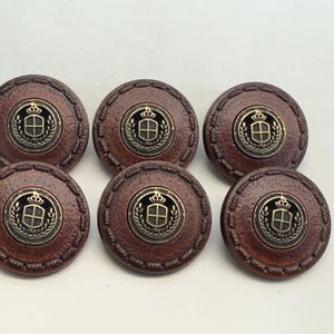 Puede incluir: Seis botones de cuero marrón con un escudo dorado que presenta un diseño de corona y escudo. Los botones tienen un borde cosido.