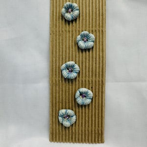 May include: A decorative piece with five ceramic flower-shaped buttons in light blue and white, with purple centres. The buttons are attached to a vertical, corrugated cardboard backing.