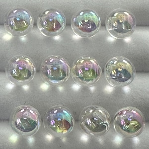 Puede incluir: Diez cuentas de vidrio iridiscentes, cada una con un brillo arcoíris. Las cuentas son redondas y lisas, y están dispuestas en tres filas sobre una superficie blanca.
