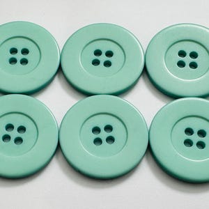 May include: Six large, round, mint-green buttons with four holes each. The buttons are arranged in two rows of three, showcasing their uniform size and color. Ideal for sewing or craft projects.