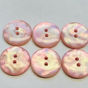 Pode incluir: Seis botões redondos rosa iridescentes, cada um com dois furos. Os botões têm um brilho perolado, com variações sutis de cor. Ideais para costura, artesanato ou para adicionar um toque decorativo às roupas.