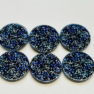 Puede incluir: Seis botones redondos con un diseño de purpurina azul oscuro y plateado. Cada botón tiene dos agujeros para coser. Los botones están colocados sobre un fondo blanco.