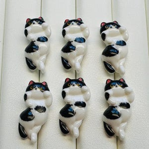 May include: Six ceramic cat figurines in a playful pose. Each cat is white with black markings, a black and red head, and yellow eyes. The figurines are arranged on a light-colored surface.