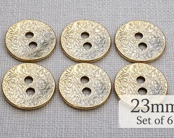 May include: Six gold-colored, round buttons with an ornate floral design and two holes. Each button is 23mm in diameter. The buttons are set on a light gray fabric.