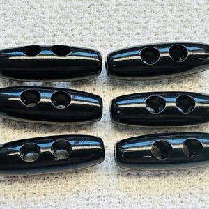 May include: Six glossy black toggle buttons with two holes each, arranged in two rows of three. The buttons have a rounded, oblong shape and are set against a textured white background.