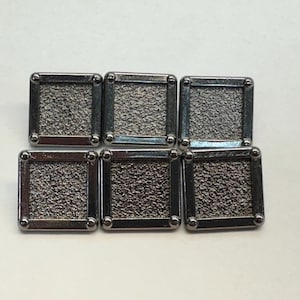 Puede incluir: Seis botones de metal plateados con una superficie texturizada y forma cuadrada.