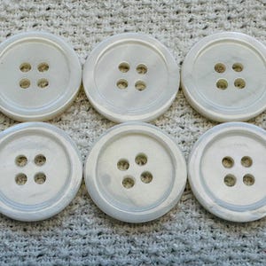 May include: Six large, round, off-white buttons with four holes each. The buttons have a subtle marbled effect and are arranged in two rows of three on a textured, light grey surface. Suitable for sewing projects.