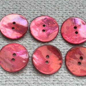 May include: Six round, iridescent red buttons with two holes each. The buttons have a pearlescent sheen, reflecting shades of pink and purple. The buttons are arranged in two rows of three.