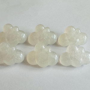 May include: Six small, cloud-shaped embellishments made of a translucent, off-white material with a fine, embedded glitter finish. Each piece features a rounded, puffy design with multiple lobes, arranged in two rows of three on a plain surface.
