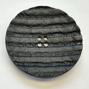Könnte beinhalten: Ein großer, runder, schwarzer Knopf mit strukturierter Oberfläche und vier kleinen Löchern. Der Knopf hat eine Reihe horizontaler, erhabener Rippen, die ein gestreiftes Muster bilden. Geeignet für Kleidung oder Bastelarbeiten.