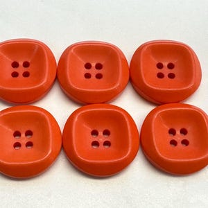 Puede incluir: Seis botones de plástico naranja con cuatro agujeros cada uno. Los botones son cuadrados con esquinas redondeadas.