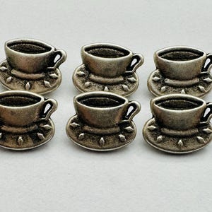May include: Six small, silver-colored teacup charms. Each charm features a teacup atop a saucer with a decorative edge. The teacups have a handle and are slightly darkened to give them an antique appearance.