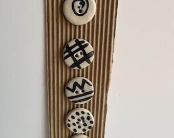 May include: A set of five off-white ceramic buttons with black abstract designs. The buttons are attached to a brown corrugated cardboard strip. Each button features a unique pattern, including swirls, crosshatch, zigzags, dots, and curved lines.