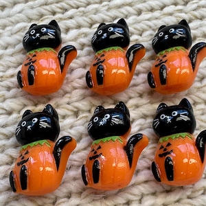 May include: Six small decorative figurines shaped like black cats sitting inside orange pumpkin bodies. Each cat has a black head with white painted eyes and whiskers, while the orange pumpkin base features a black jack-o'-lantern face design.