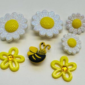 May include: A collection of decorative embellishments featuring daisies and a bee. The daisies have white petals with iridescent glitter and yellow centers. Two yellow flower-shaped embellishments with a gem center are also included.