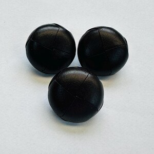 Puede incluir: Tres botones negros y redondos con apariencia cosida y texturizada. Los botones están dispuestos en un patrón triangular sobre un fondo blanco. Estos botones son adecuados para proyectos de costura y artesanía.