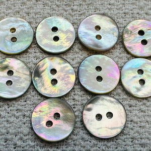 May include: Ten round, iridescent buttons with two holes each. The buttons have a pearlescent sheen with hints of pink, green, and yellow. They are arranged on a textured, light gray surface.