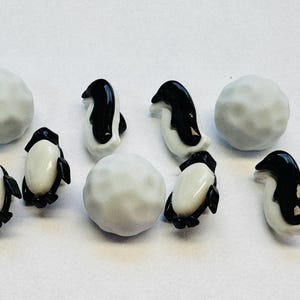 May include: A collection of miniature penguin figurines and small, round, gray objects resembling snowballs. The penguins are black and white, with a glossy finish. The items are arranged on a white surface.