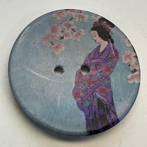 Könnte beinhalten: Runder Holzknopf mit einem bemalten Design. Der Knopf zeigt eine Frau in einem lila Kimono unter einem rosa blühenden Baum. Der Hintergrund ist hellblau. Er hat zwei Löcher zum Nähen.