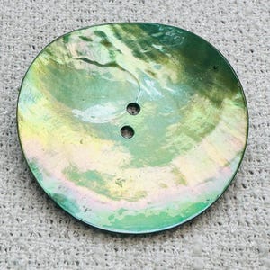 May include: A large, round button with a pearlescent finish. The button has a green and gold iridescent sheen, with two small holes near the center. The button is set against a textured, off-white background.