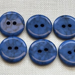 Puede incluir: Un conjunto de seis botones redondos de dos agujeros con un acabado liso y brillante con un patrón jaspeado en azul y gris oscuro. Cada botón tiene una superficie ligeramente abovedada y una forma circular consistente, dispuestos en dos filas.