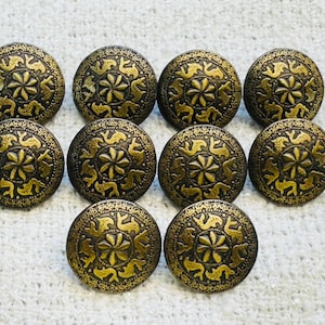 Puede incluir: Diez botones decorativos de color latón antiguo. Cada botón tiene un diseño circular con un motivo floral central rodeado de figuras de animales estilizadas. Los botones están dispuestos sobre una superficie blanca texturizada.